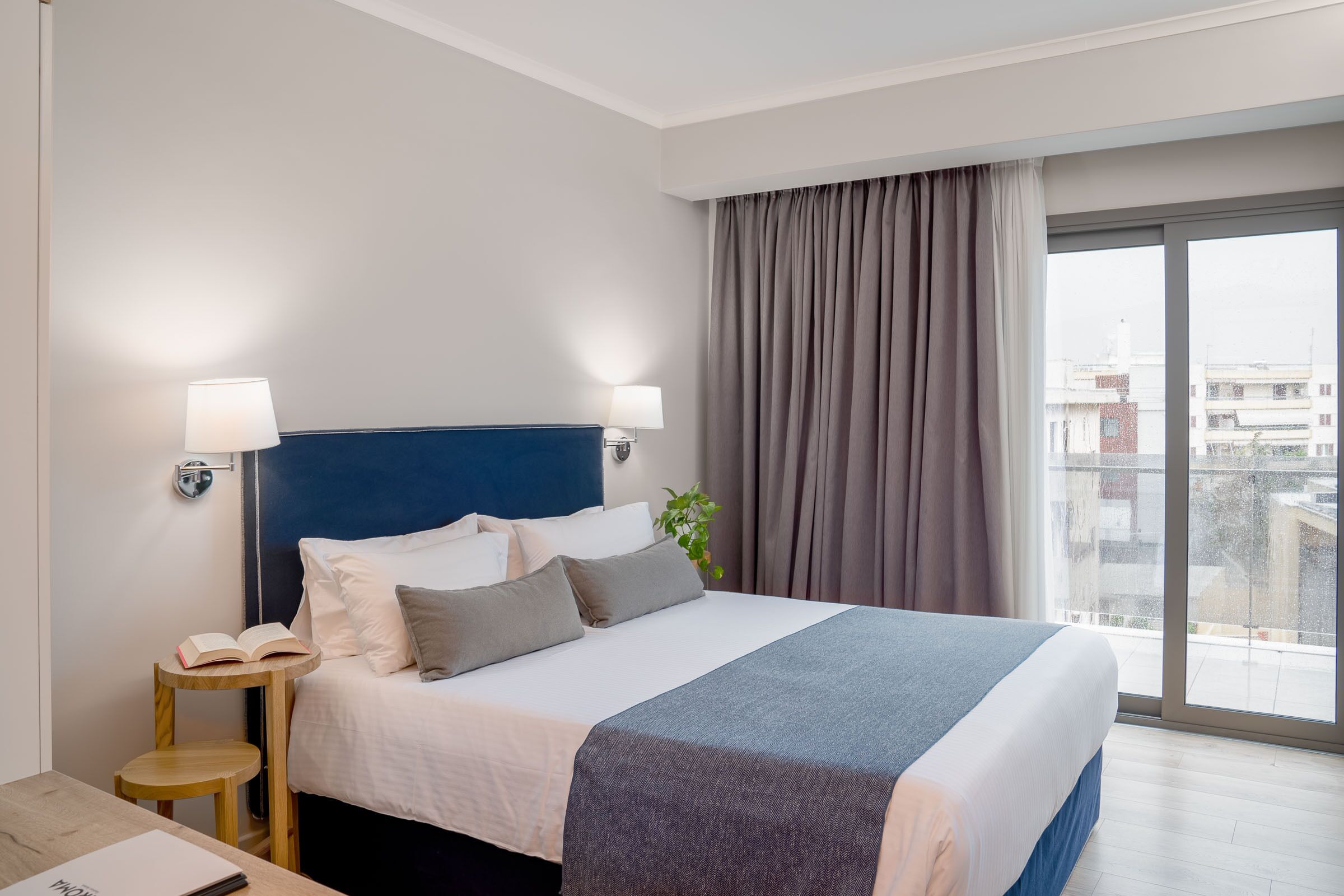 A hotel room with a large bed, two bedside lamps, a small wooden table with a book, and a view of the city through a large window.