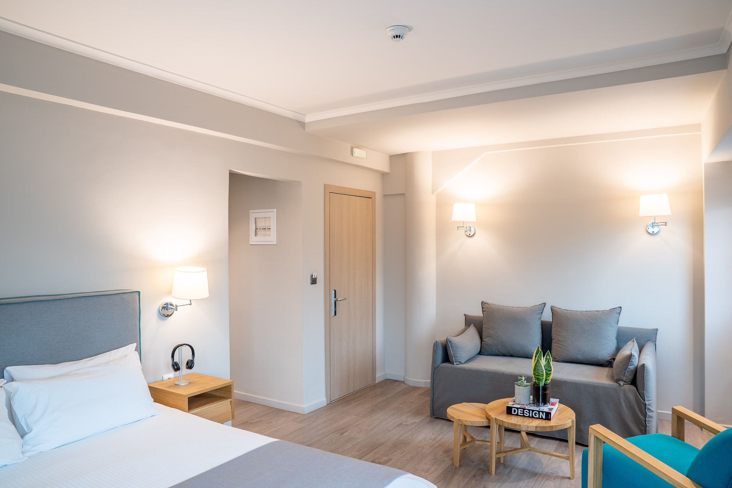 Hotellihuone, jossa on sänky, sohva, pieni pöytä ja sen päällä kirja sekä kaksi seinälle asennettua lamppua.