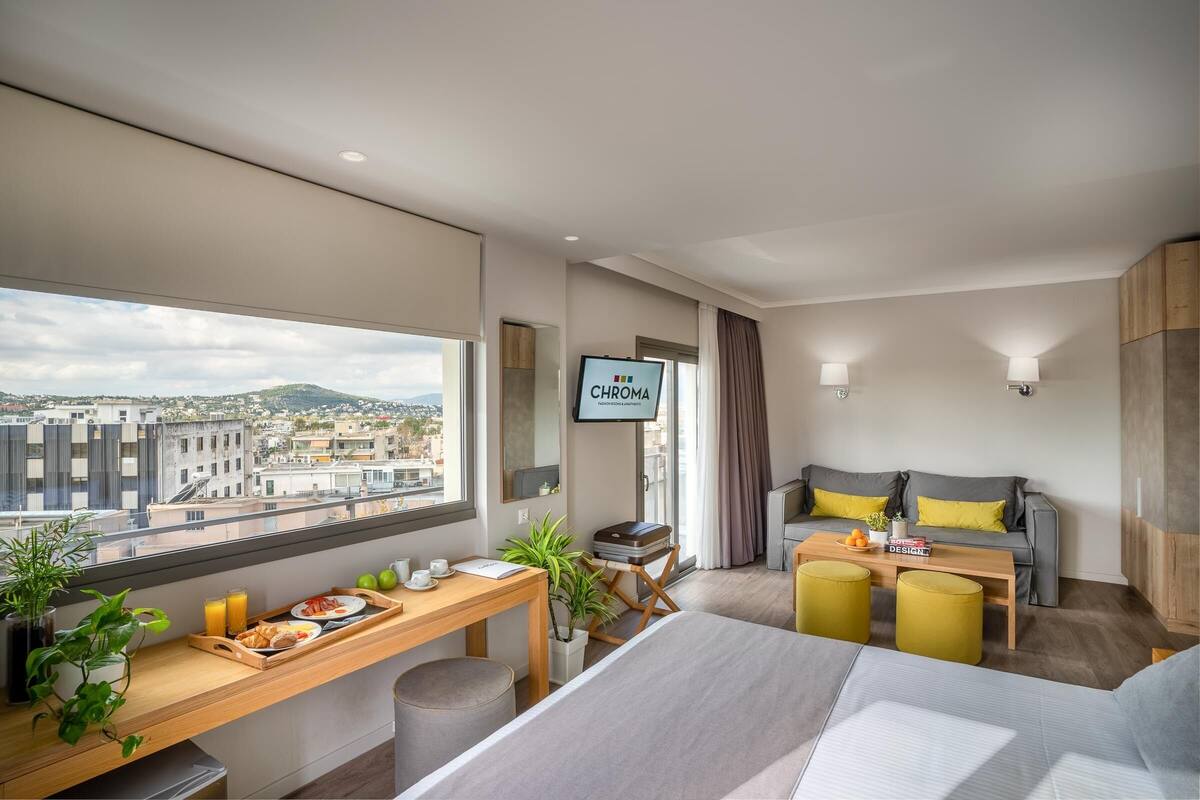 A modern hotel room with a city view, a desk with breakfast items, a sofa, and a television.