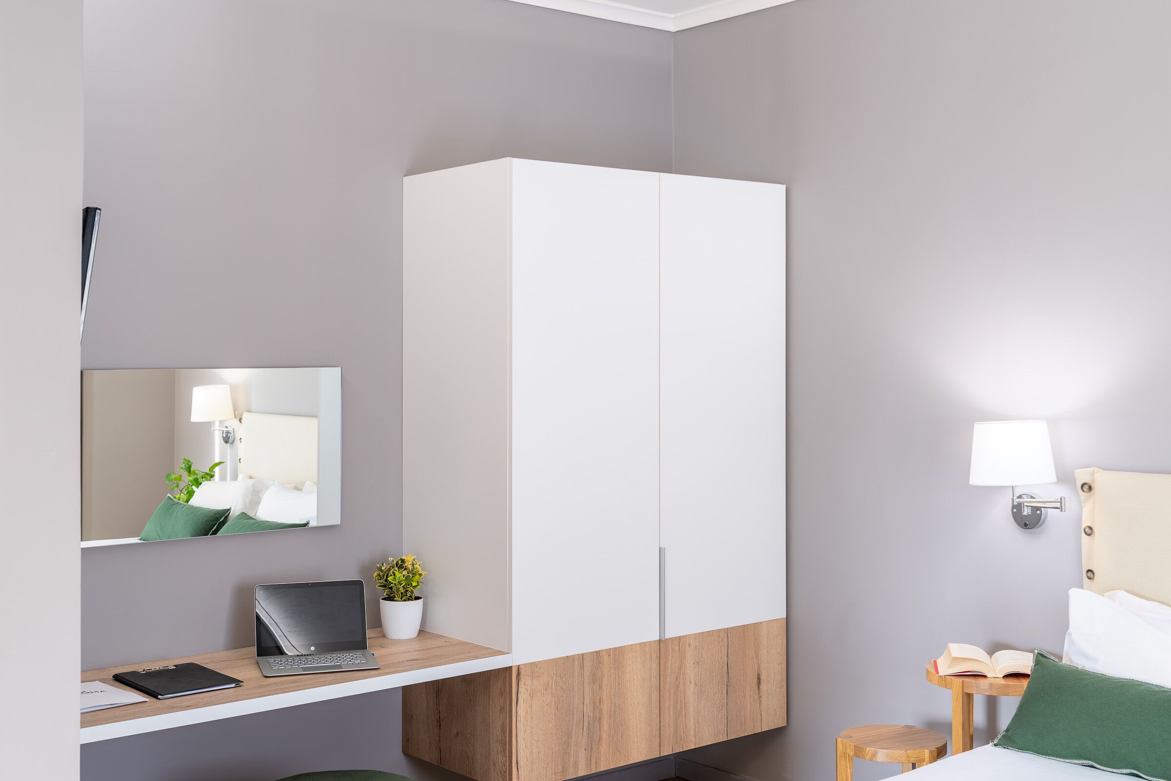 Una habitación de hotel moderna con un escritorio de madera, una laptop, una planta pequeña en maceta, una cama con almohada verde y una mesita de noche con una lámpara.