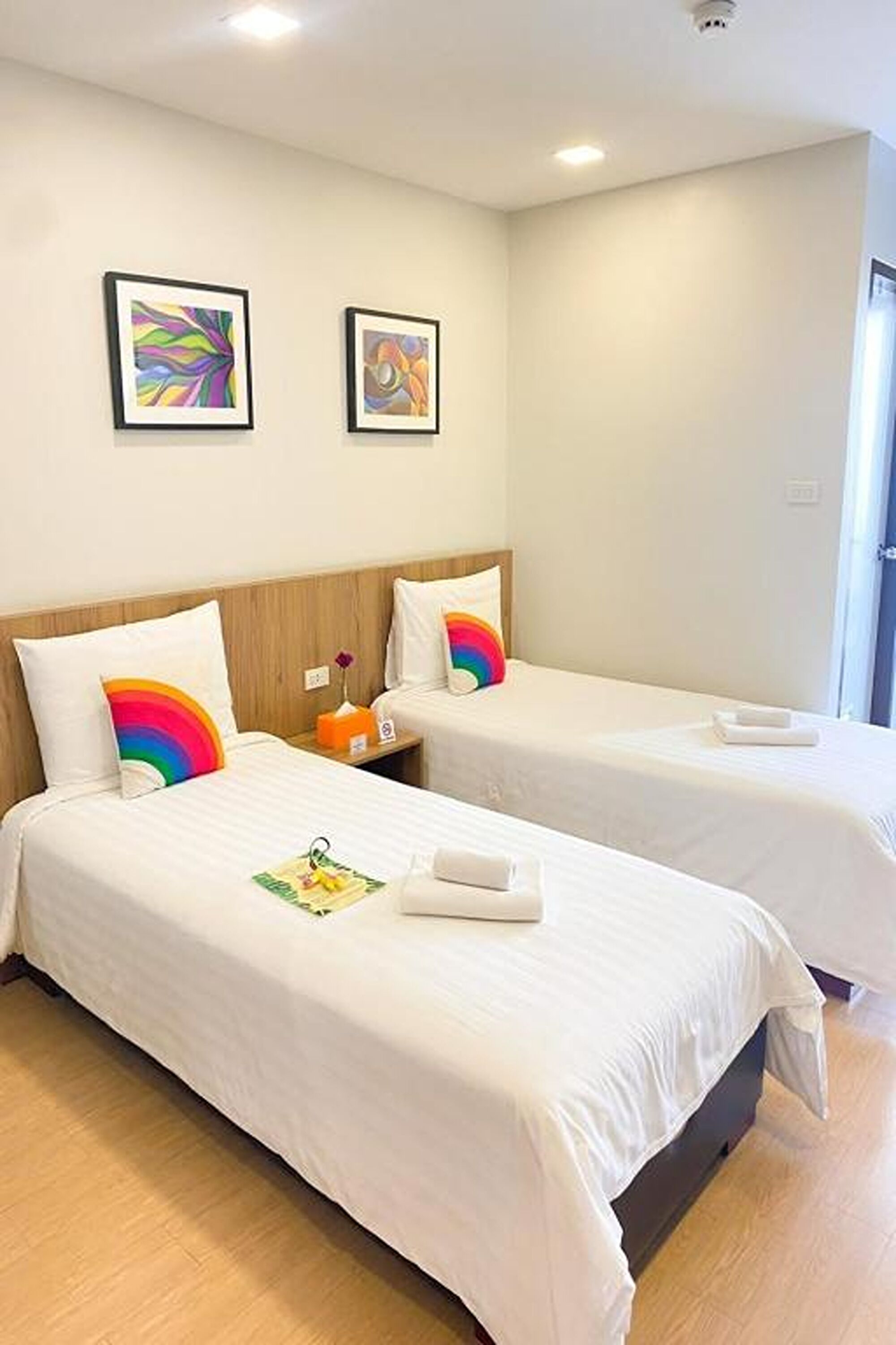 A hotel room with two single beds, white bedding, and colorful pillows. There are framed artworks on the wall and a small table with a lamp.