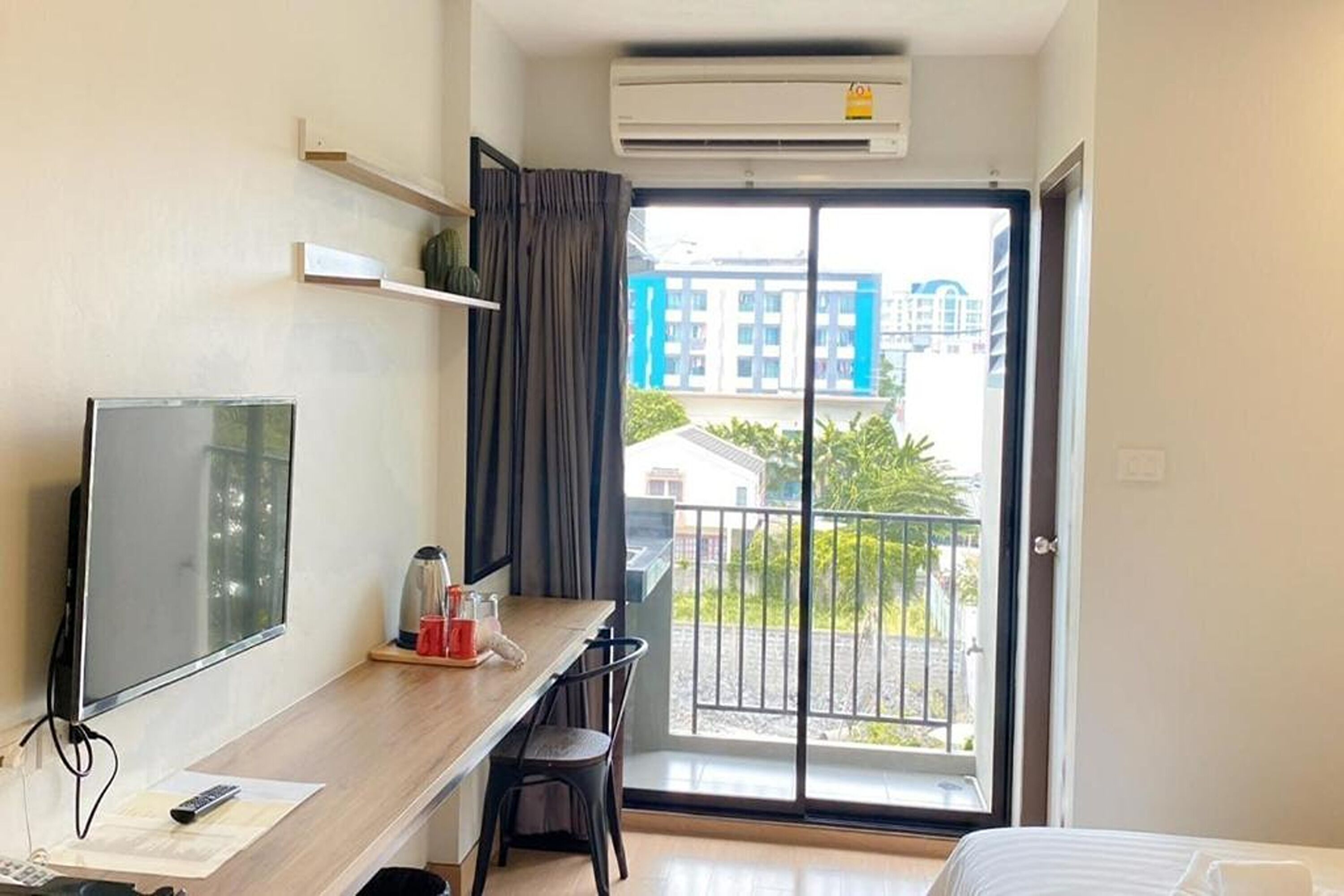 A hotel room with a bed, a desk with a TV, a chair, a window with a view of buildings, and an air conditioning unit.