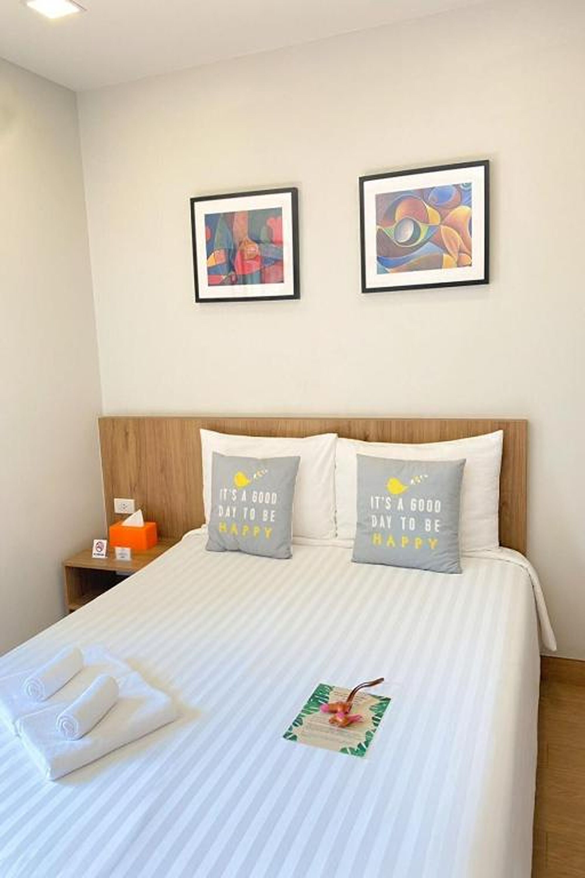 A neatly made bed with striped bedding, two pillows with motivational quotes, a framed abstract painting above the bed, and a small nightstand with a tissue box and a sign.