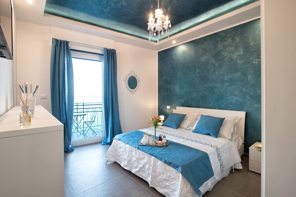 Quarto com cama, mesa de cabeceira, cômoda, lustre, janela com cortinas e varanda.