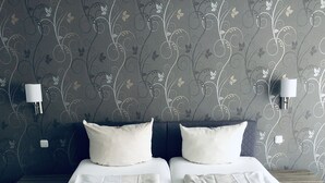 A double bed with white linens and two pillows, a bedside table, and a wall with a floral pattern. - Der Watzenhof (Hemsbach)
