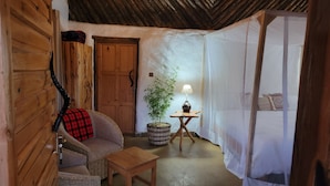 A cozy room with a wooden door, a wicker sofa with a red and black checkered throw, a small wooden table, a bamboo plant, and a table lamp. - Maji Moto Maasai Cultural Camp (Maji Moto)