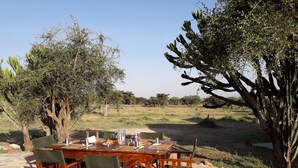 An outdoor dining area with a wooden table and chairs, surrounded by trees and a grassy landscape. - Maji Moto Maasai Cultural Camp (Maji Moto)