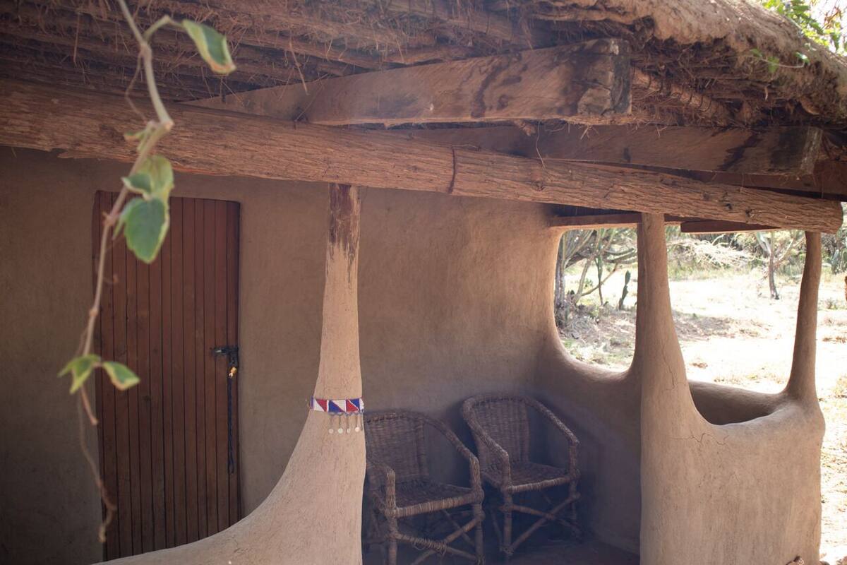 A rustic hut with a wooden door, two wicker chairs, and a thatched roof.