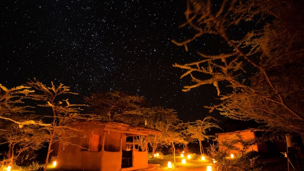 A small, rustic building with a thatched roof, surrounded by trees and illuminated by numerous ground lights. - Maji Moto Maasai Cultural Camp (Maji Moto)