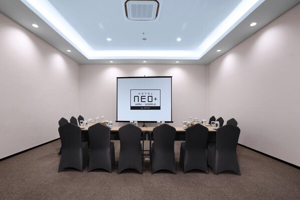 A conference room with a long table, black chairs, a projector screen, and a ceiling light fixture.