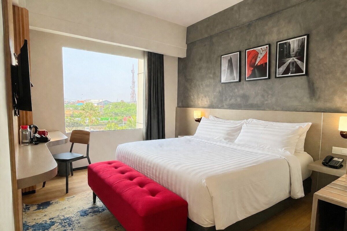 Habitación de hotel con cama, un banco rojo, un escritorio con silla, televisor y ventana con vistas.