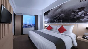 Habitación de hotel con una cama grande, un televisor de pantalla plana montado en la pared, vista panorámica de la ciudad a través de una ventana y una obra de arte grande con marco sobre la cama.