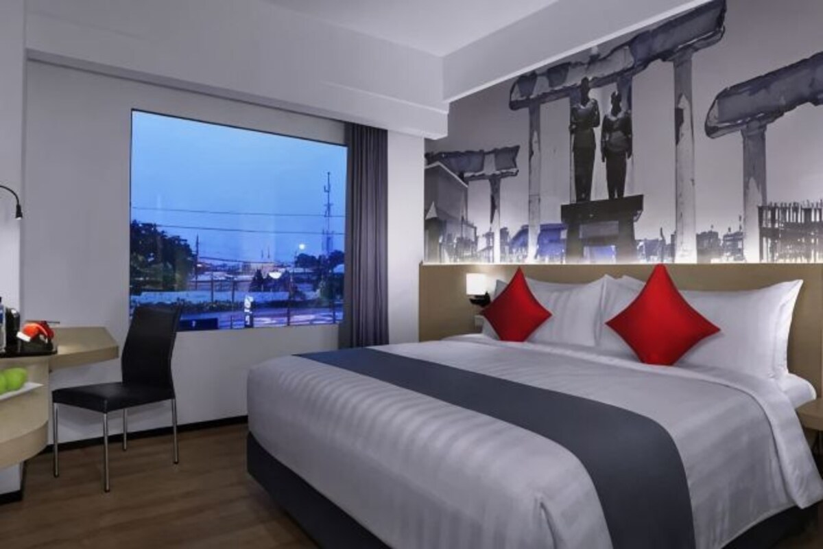 A hotel room with a large bed, a desk, and a view of a cityscape through a large window.