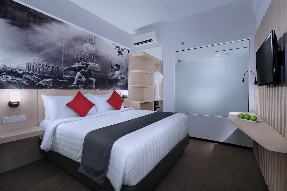 Moderni hotellihuone, jossa on suuri sänky, taulutelevisio, vaatekaappi ja seinälle kiinnitetty hylly, jossa on hedelmiä.