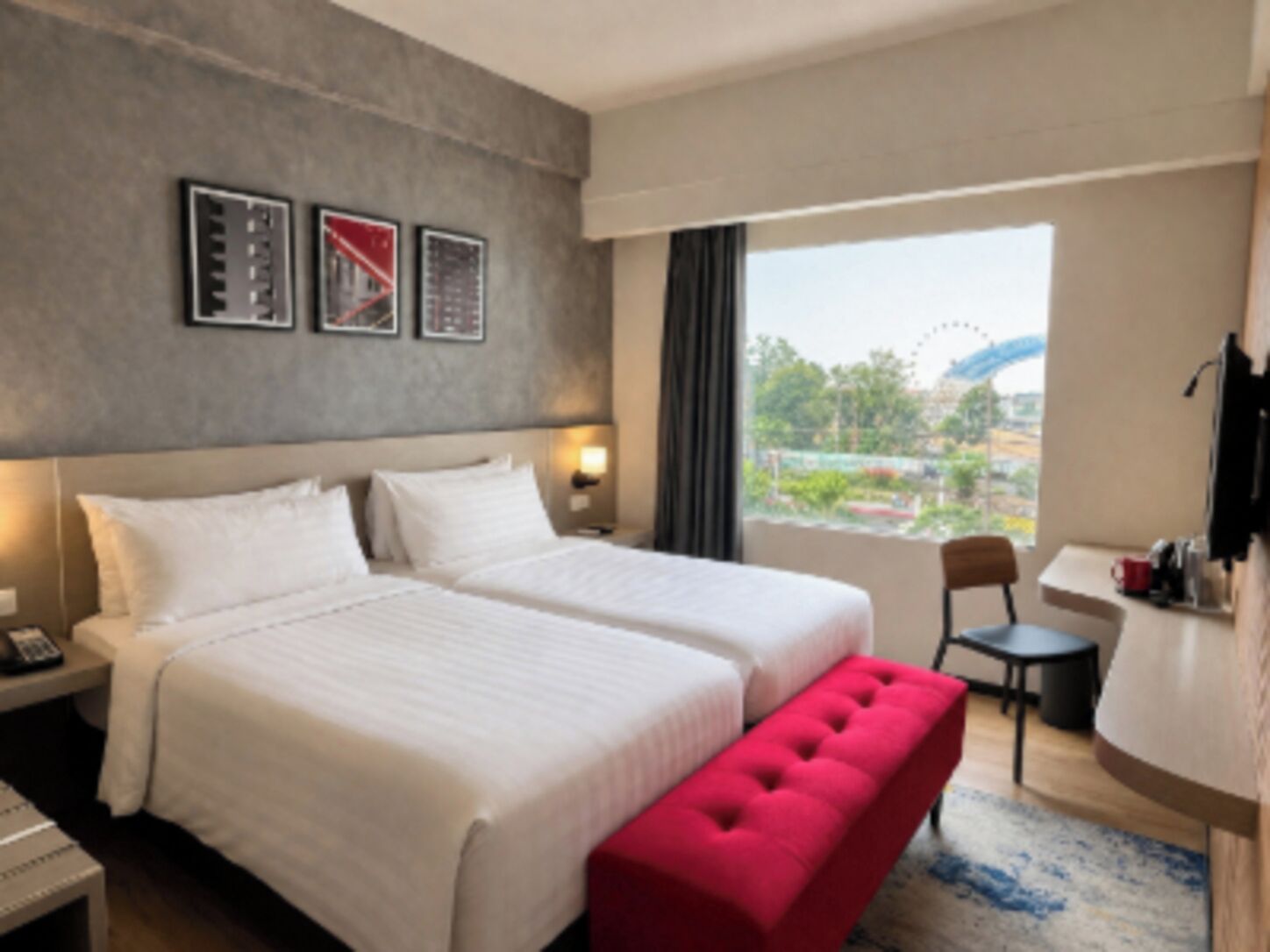 A hotel room with two beds, a red bench, a desk, and a view of a cityscape.