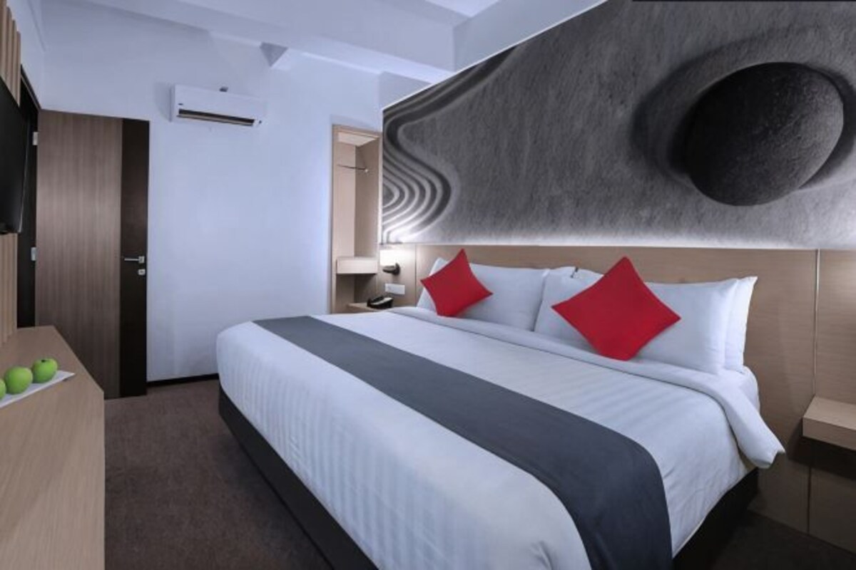 A modern hotel room with a large bed, a flat-screen TV, a desk with apples, and a unique wall design.