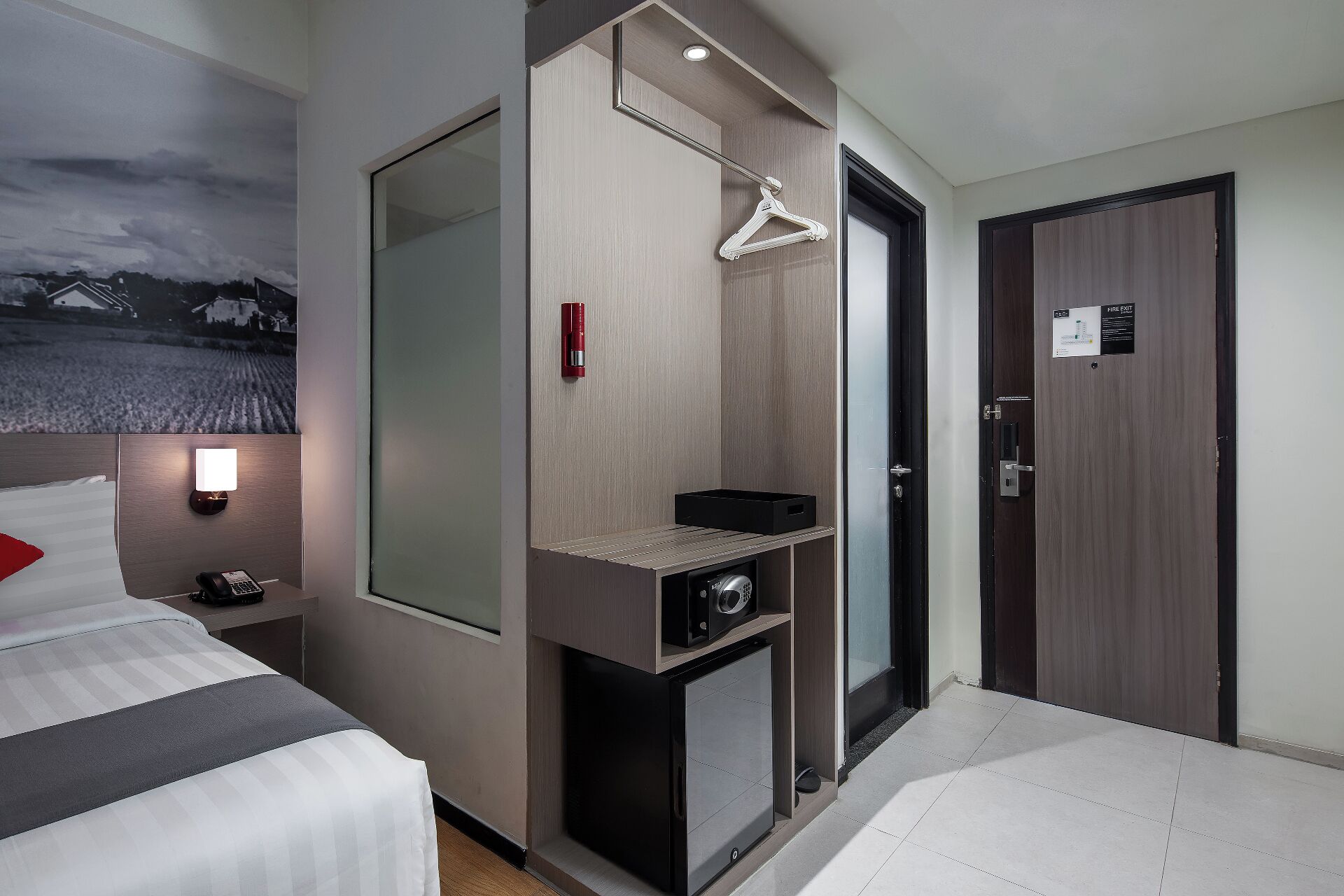 A hotel room with a bed, a desk with a computer, a mirror, a fire exit sign, and a view of a landscape.