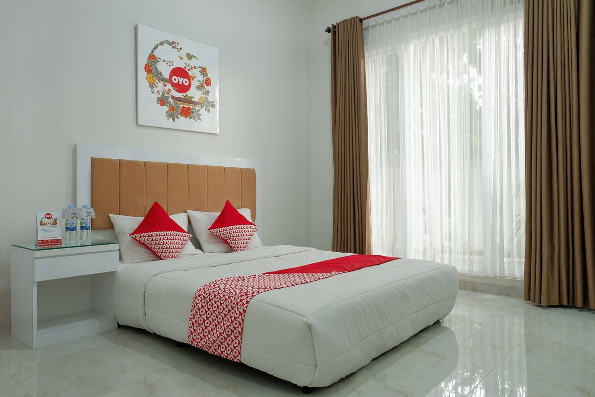A neatly made bed with a white and red patterned bedspread, a wooden headboard, and two red pillows. There is a white bedside table with a glass top and a few items on it. A framed picture hangs on the wall above the bed.