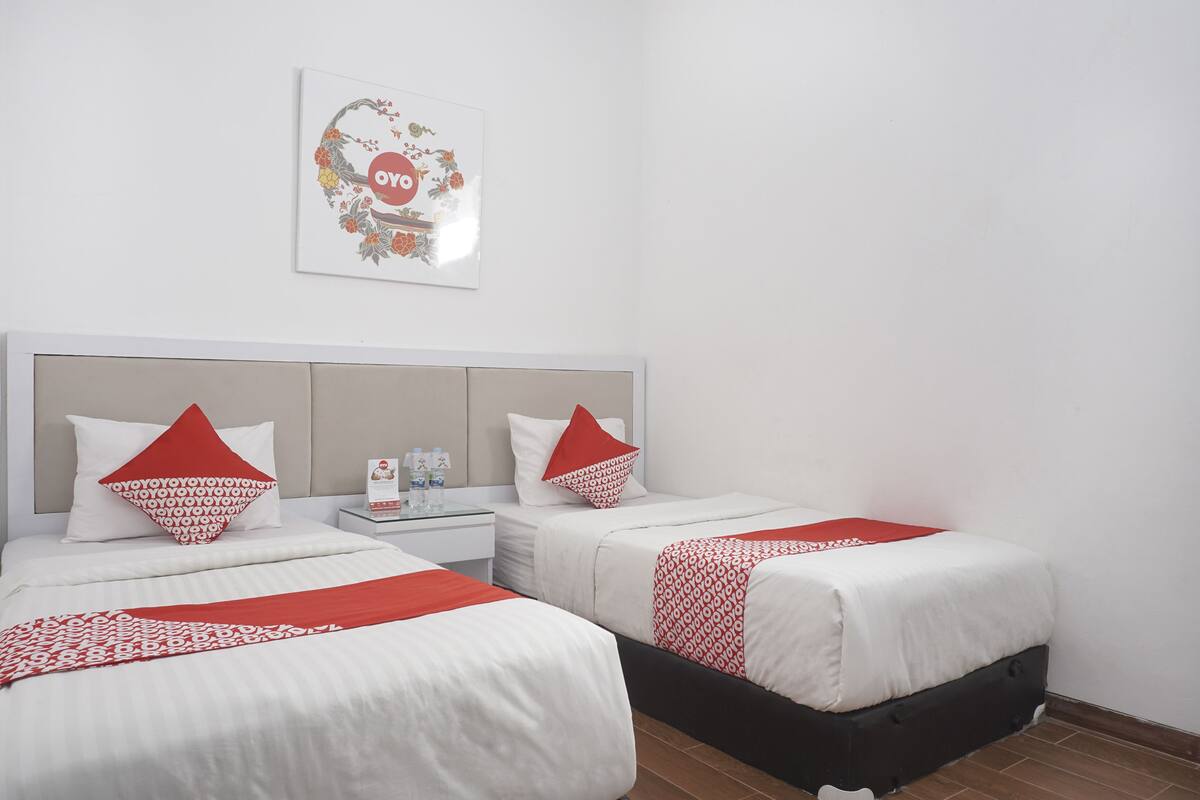 A hotel room with two beds, each with white bedding and red accents, a nightstand with toiletries, and a framed artwork on the wall.