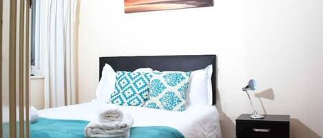 Een opgemaakt bed met gestreepte kussens, een bedlampje en een aan de muur hangend schilderij.