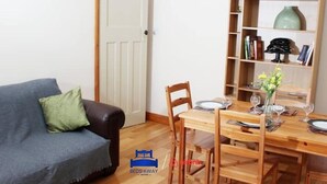 Une pièce chaleureuse avec une table à manger dressée pour quatre personnes, une bibliothèque remplie de divers objets et un canapé gris agrémenté d’un coussin vert.