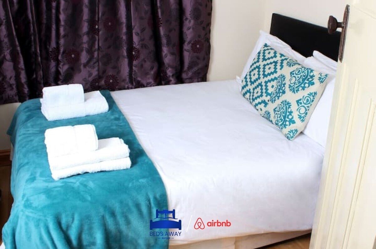 A neatly made bed with white linens, a teal throw blanket, and a patterned pillow. There are two folded white towels on the bed.