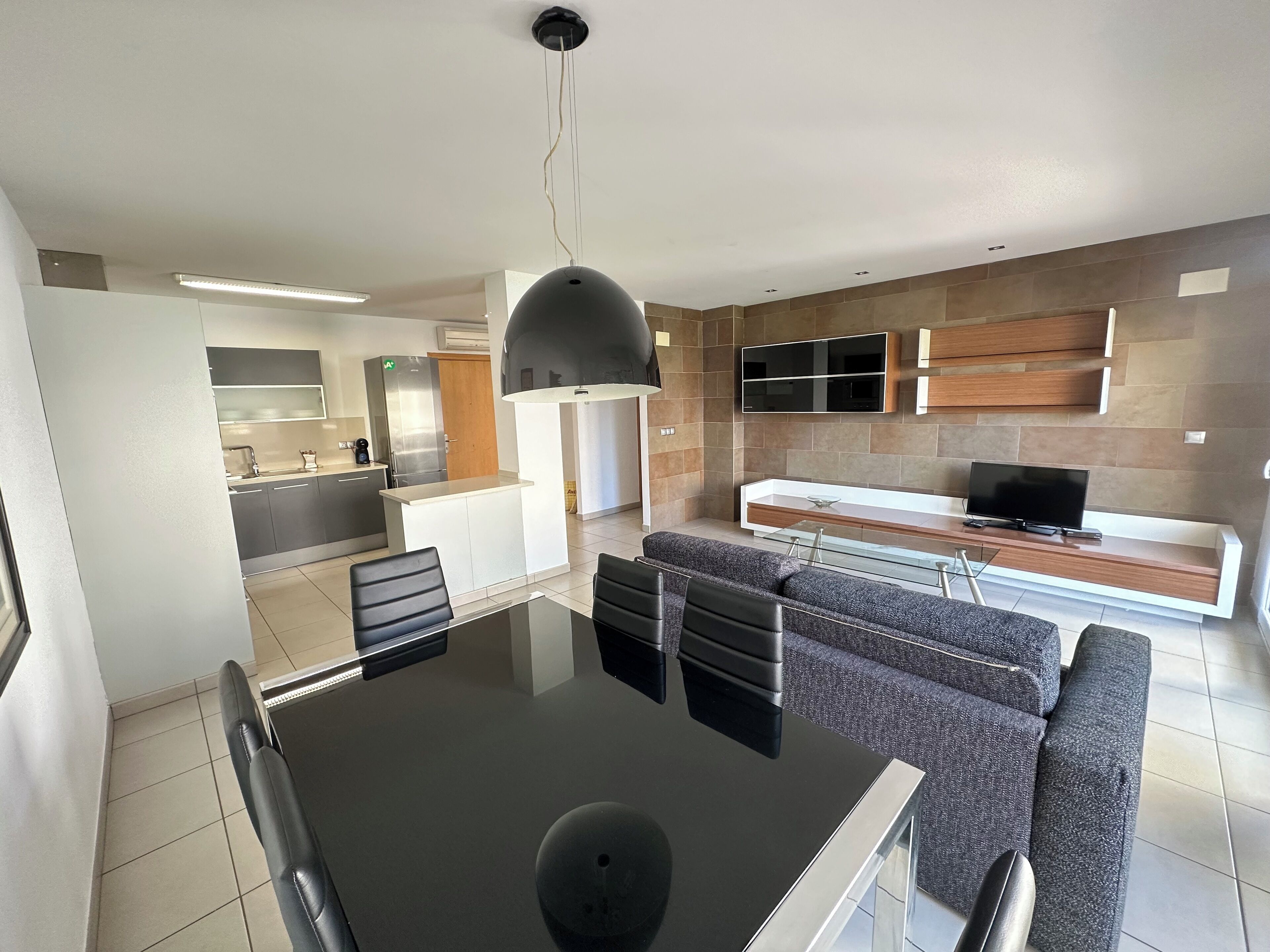 Een moderne keuken met een centraal eiland, een eetruimte met een glazen tafel en stoelen, en een zithoek met een bank en televisie.