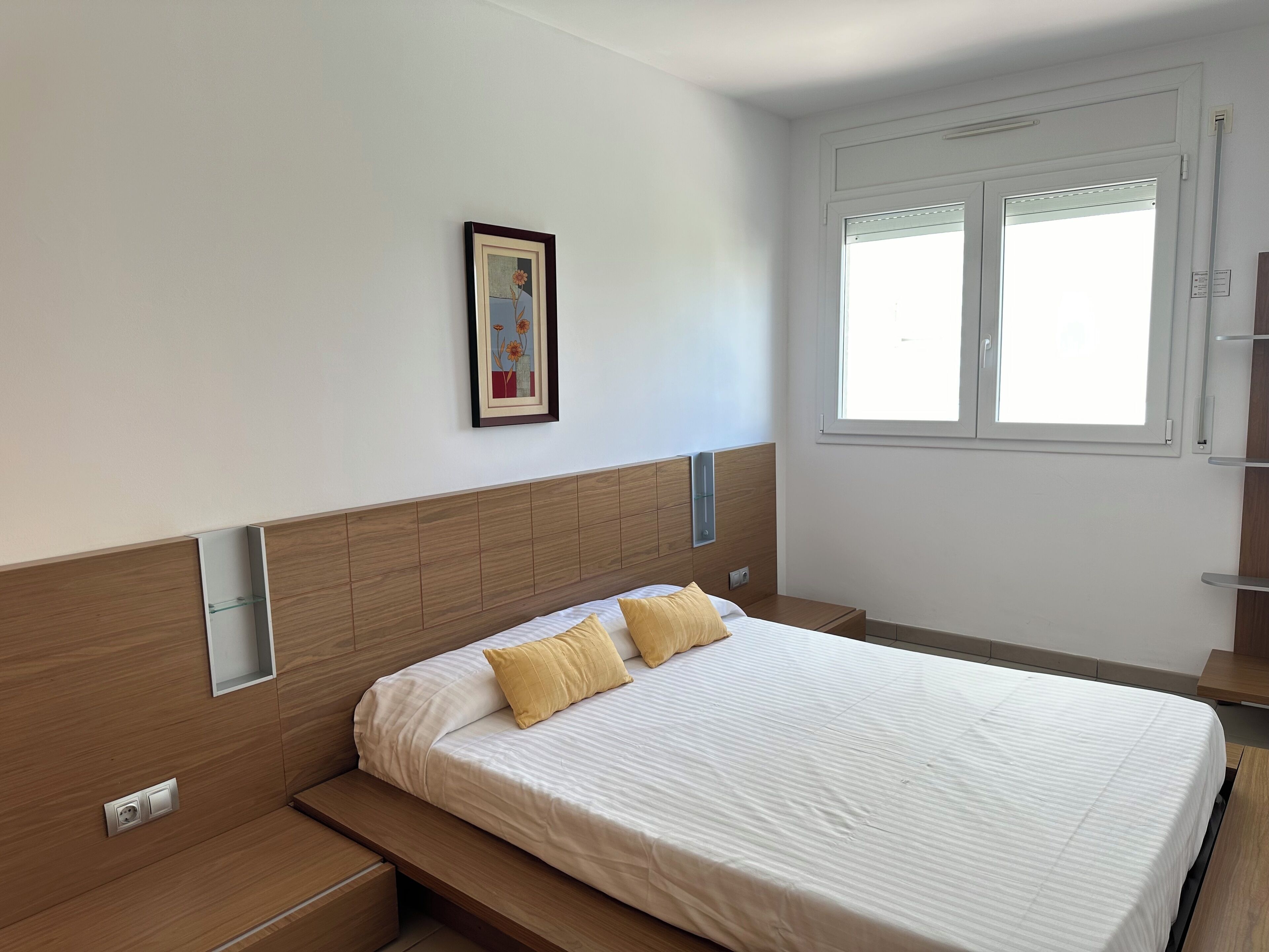 A modern bedroom with a wooden headboard, a bed with white bedding and two yellow pillows, a bedside table, and a window with blinds.