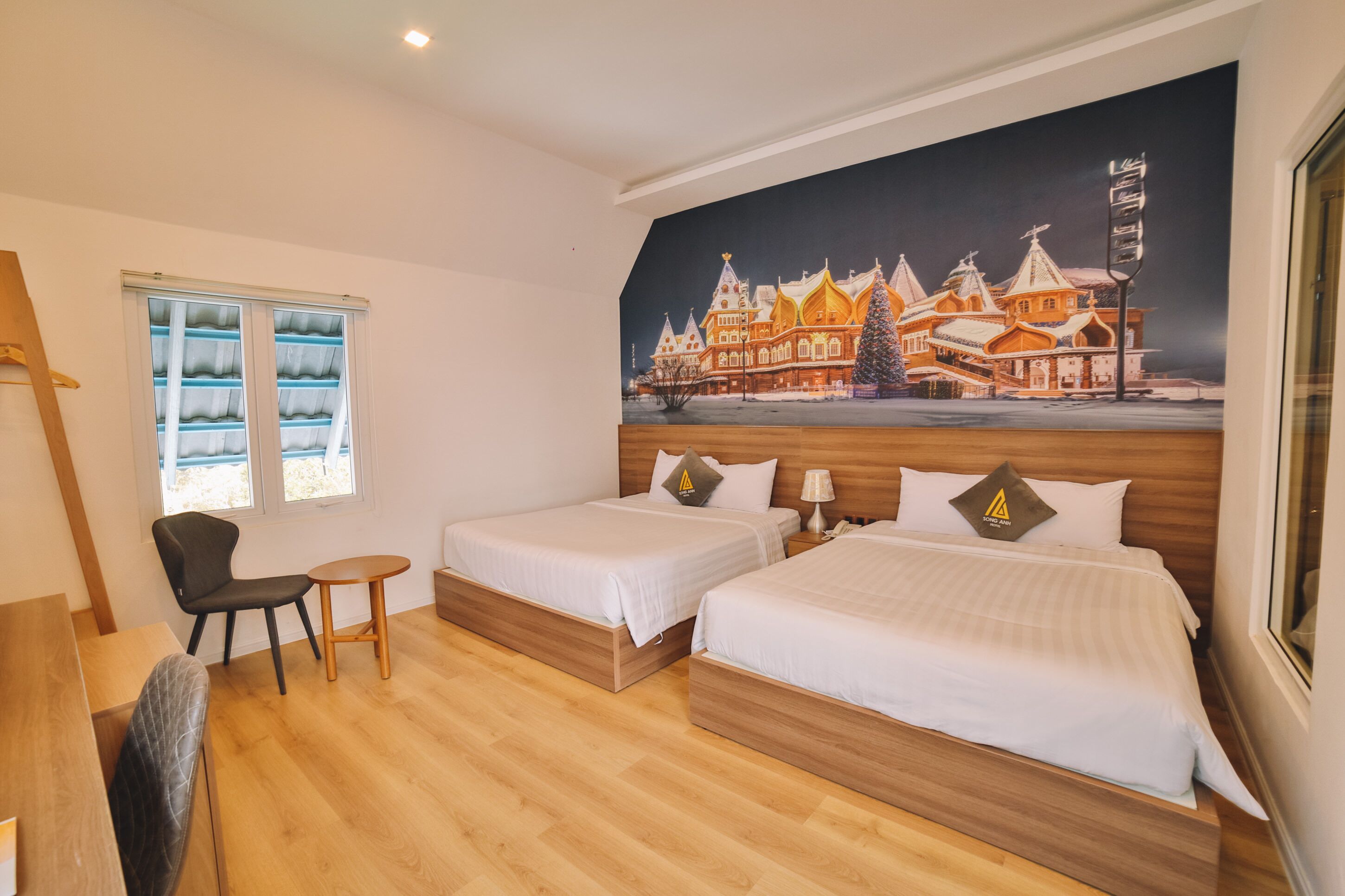 A hotel room with two beds, a wooden floor, a window with blinds, a small table, and a chair.