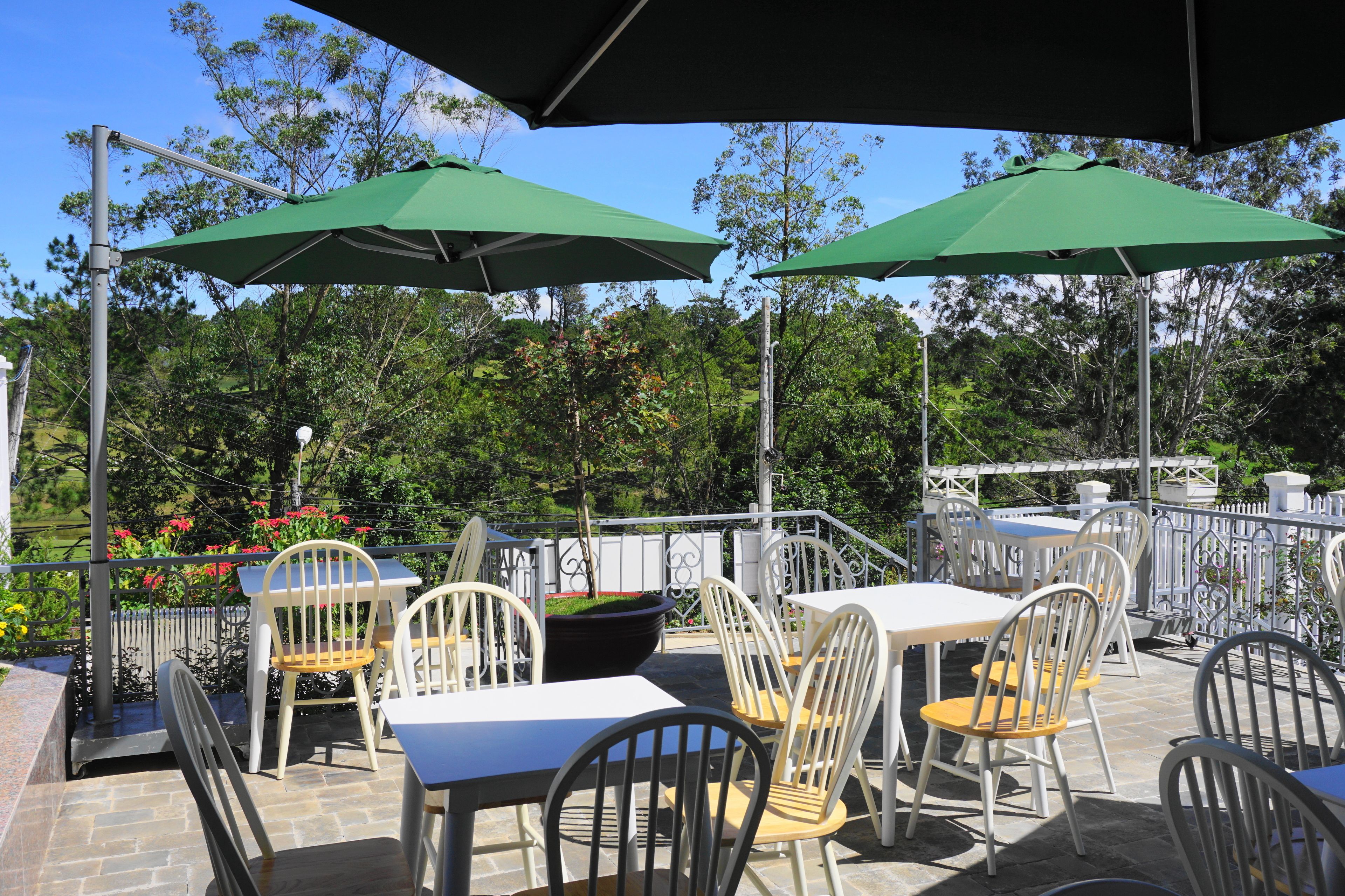 Un espace extérieur aménagé en terrasse, avec des tables et des chaises, des parasols verts et une vue sur les arbres et les fleurs.