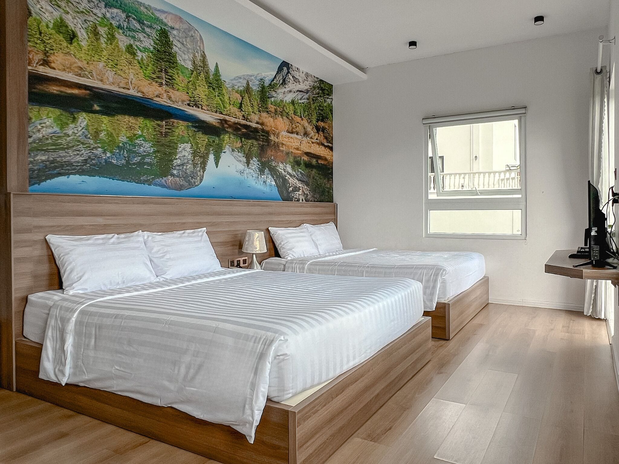 A modern hotel room with two beds, a large scenic painting, a window with a view, and a desk with a computer.