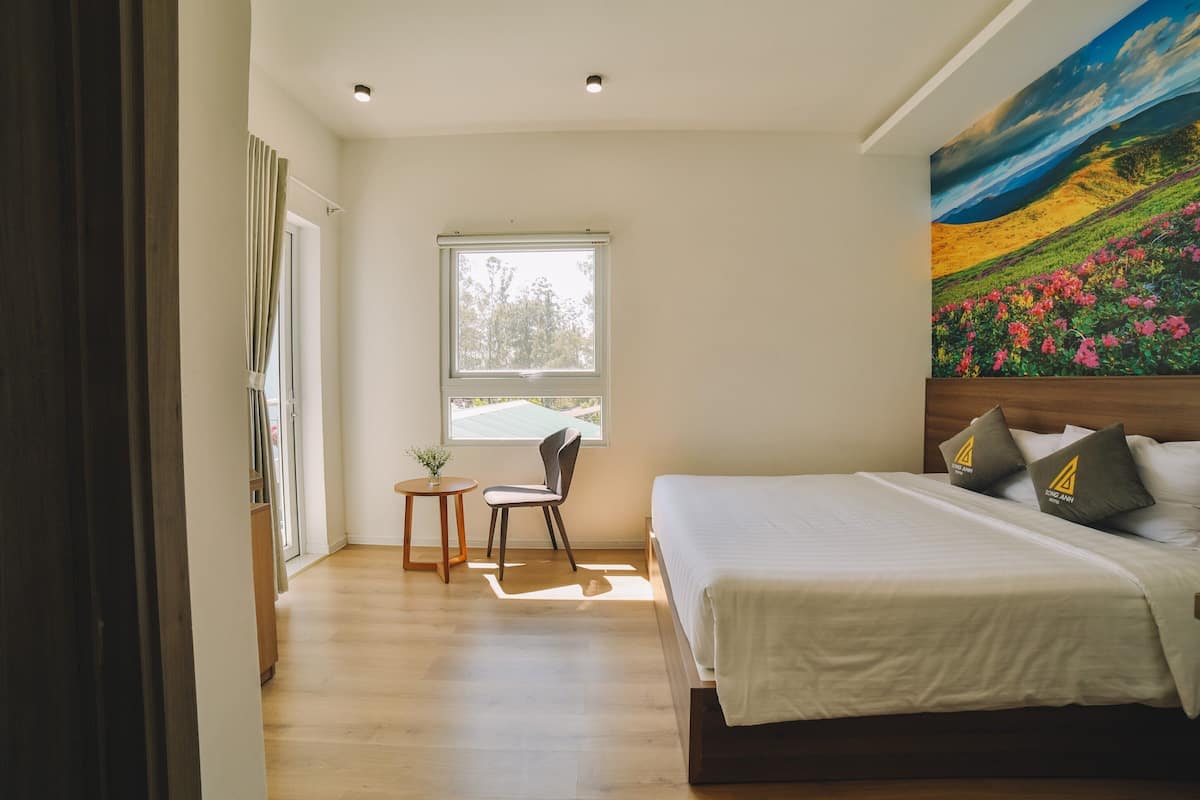 A bedroom with a large bed, a small table, a chair, a window, and a painting on the wall.