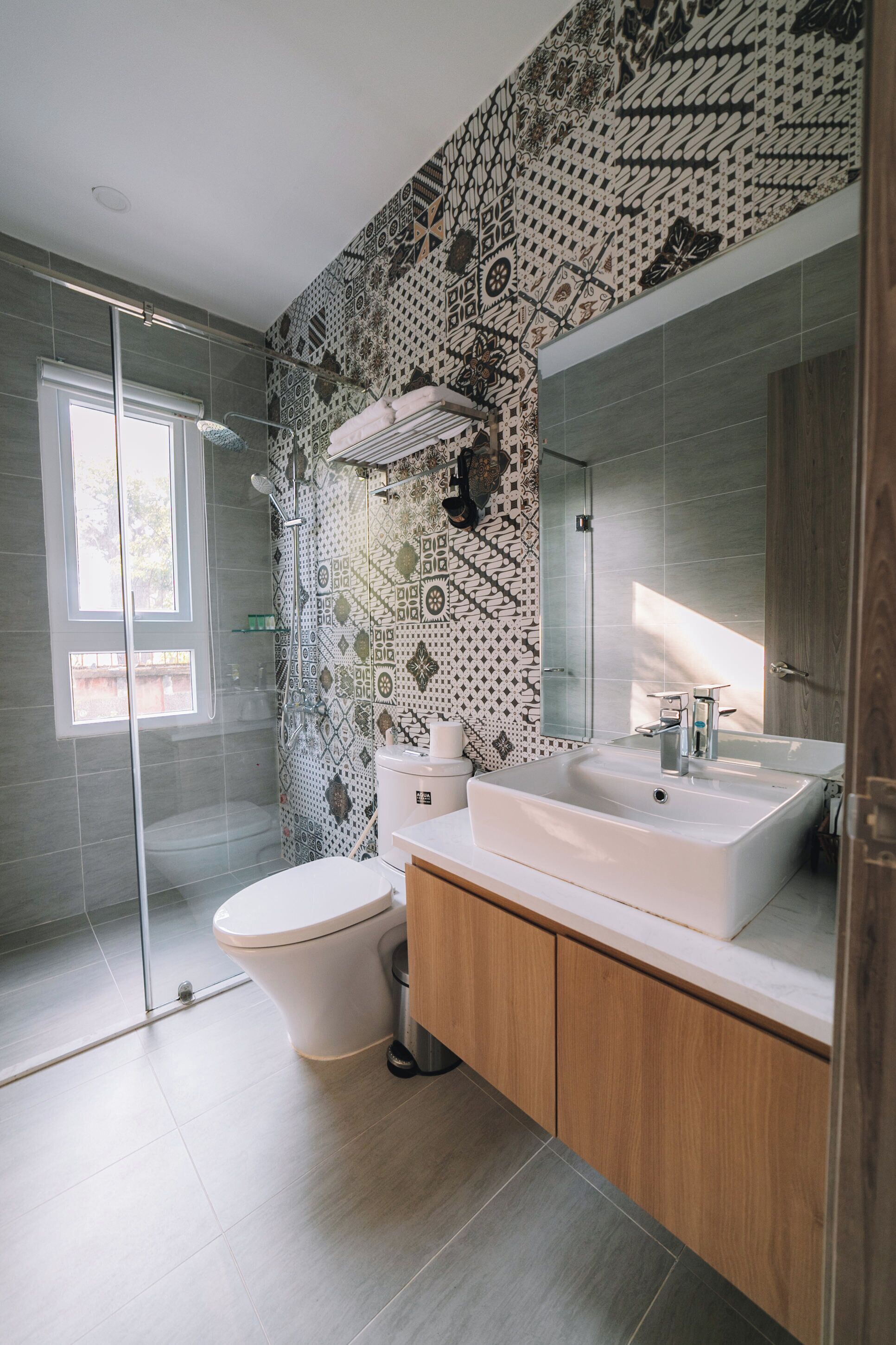 A modern bathroom with a patterned wall, a glass shower enclosure, a white toilet, and a wooden vanity with a sink.