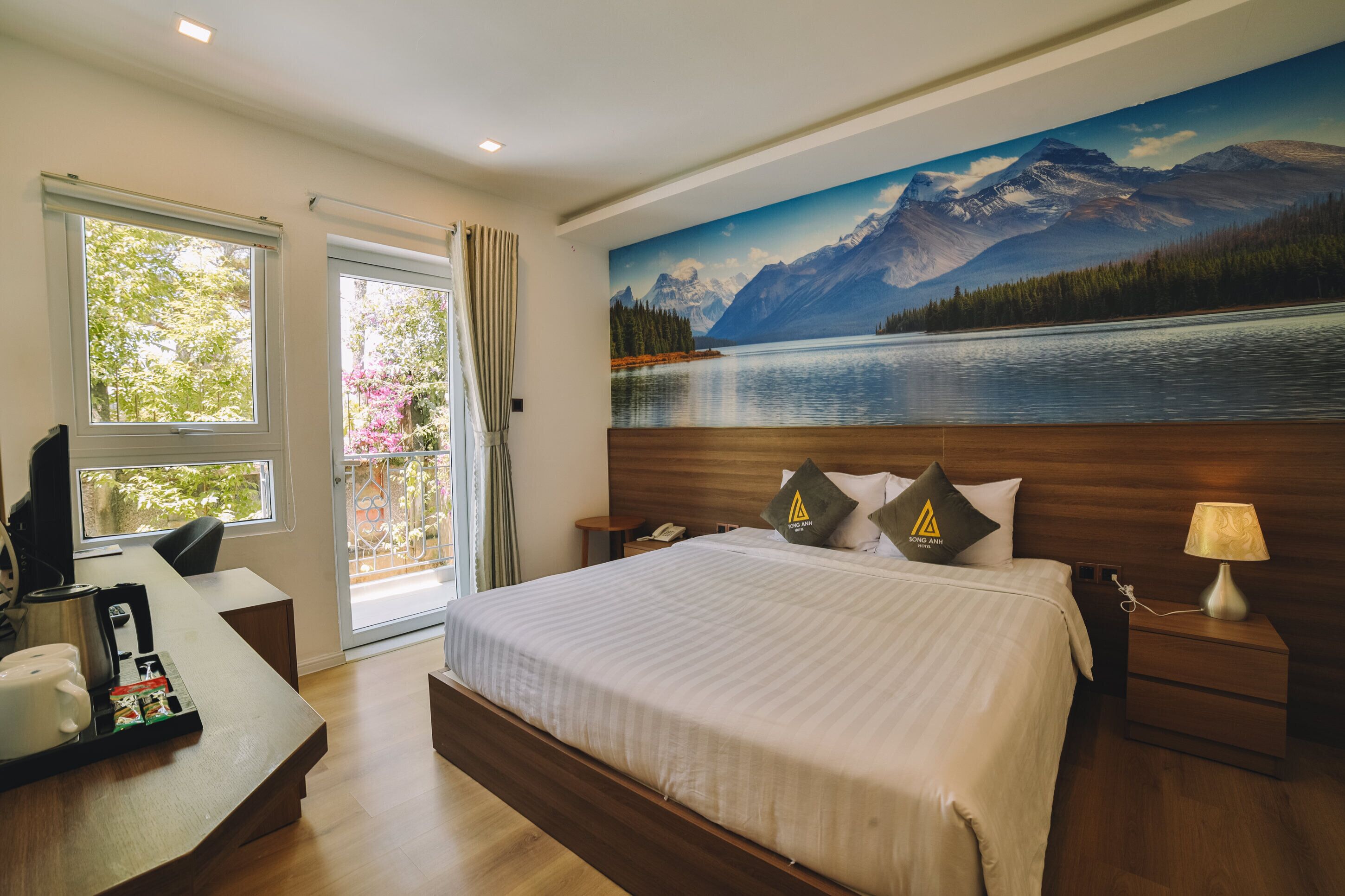 Habitación de hotel con una cama grande, un escritorio con cafetera, un cuadro de un paisaje montañoso y vistas a un lago.