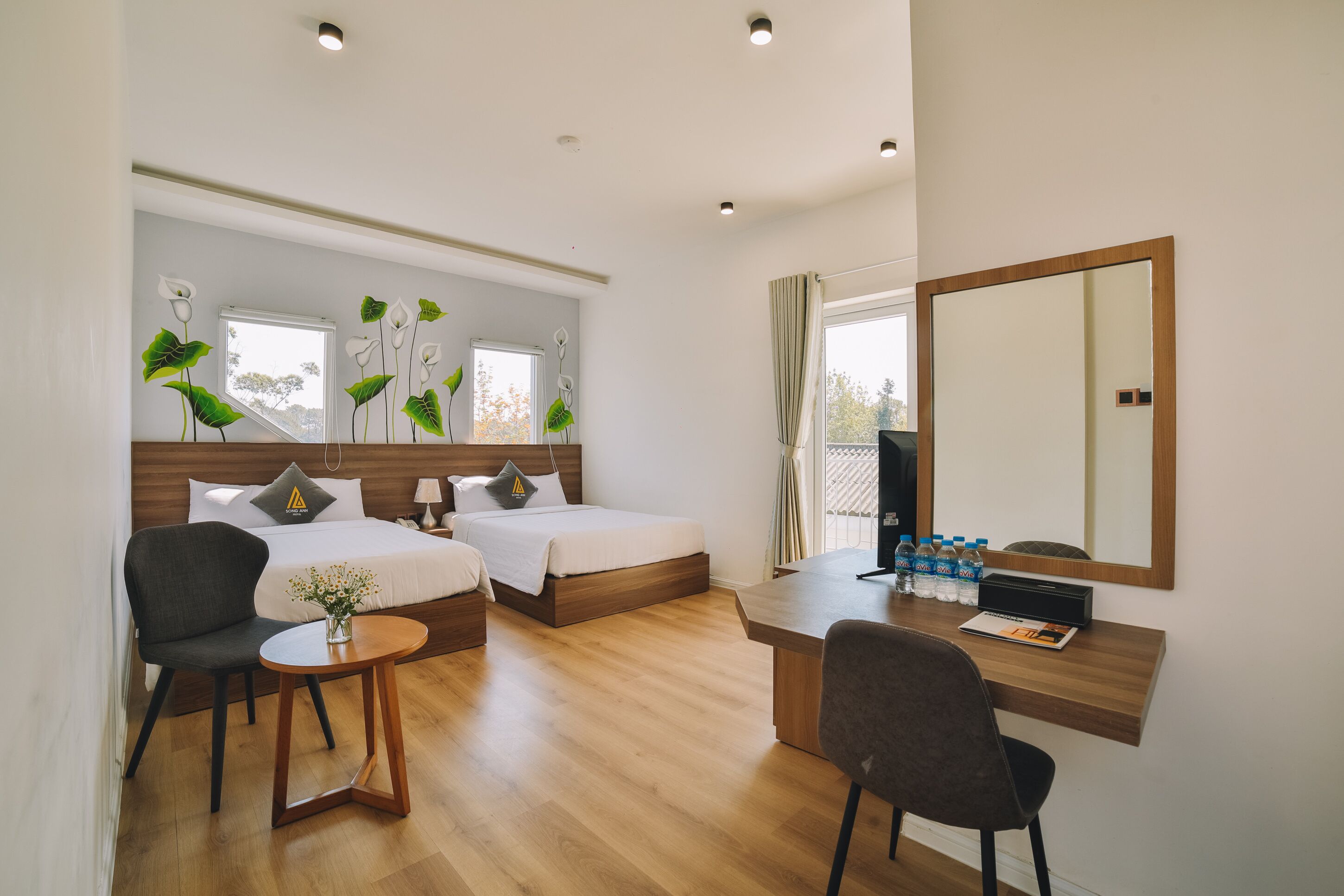 A hotel room with two beds, a desk, a chair, a small table with a vase, and a large mirror.