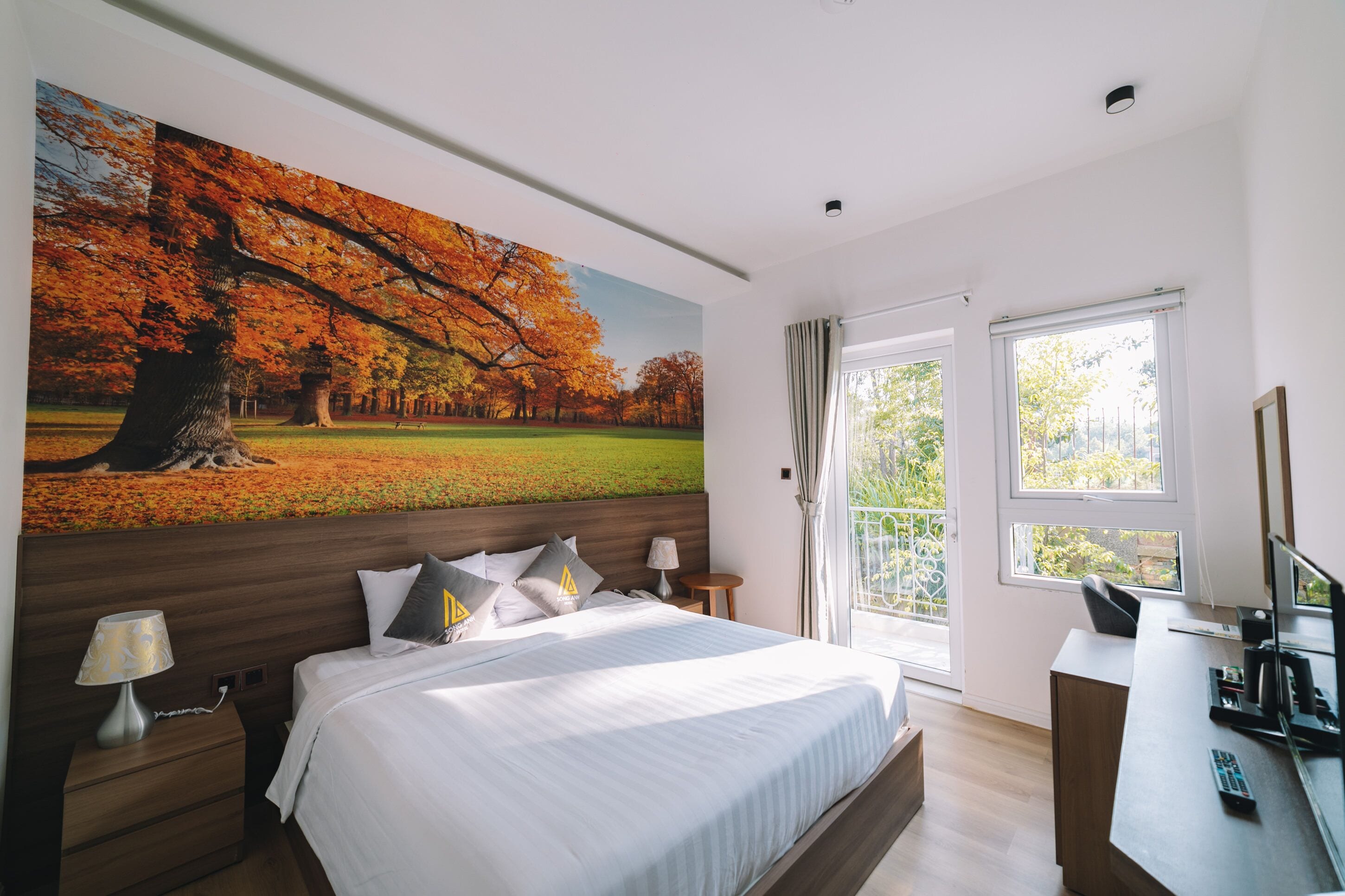 Une chambre à coucher avec un grand lit, un bureau, une chaise, une lampe et un tableau représentant des arbres d'automne.