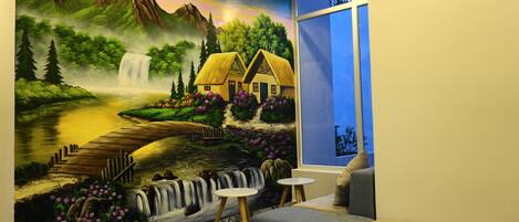 Habitación con un mural de un paisaje que muestra una casa, una cascada y un río. Se ve una ventana con vistas al exterior, un sofá y una mesita.