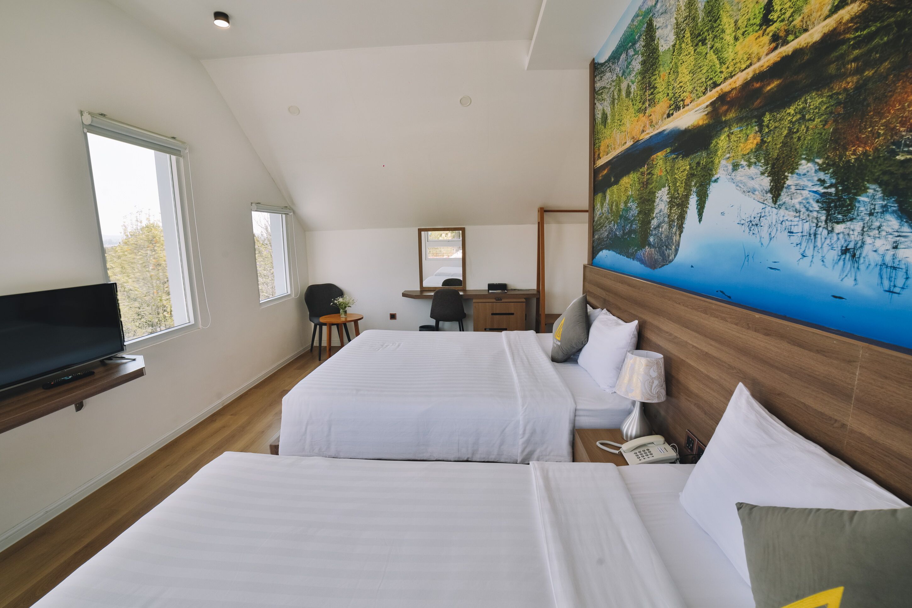 Une chambre d’hôtel avec deux lits, un bureau, une chaise, une télévision et un grand tableau représentant un lac.