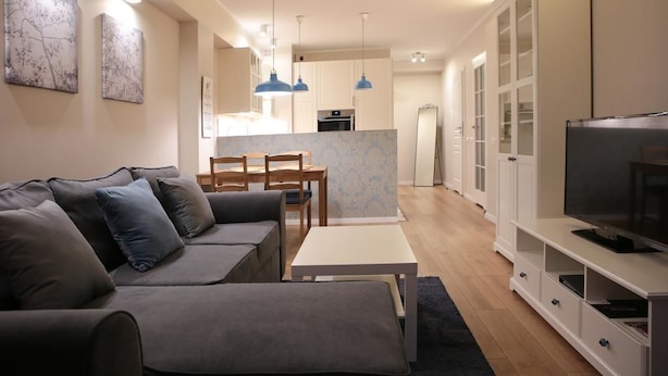 Een moderne woonkamer met een grijze bank, een klein tafeltje en een televisie op een witte kast. Achtergrond is een keuken met kasten en een koelkast.