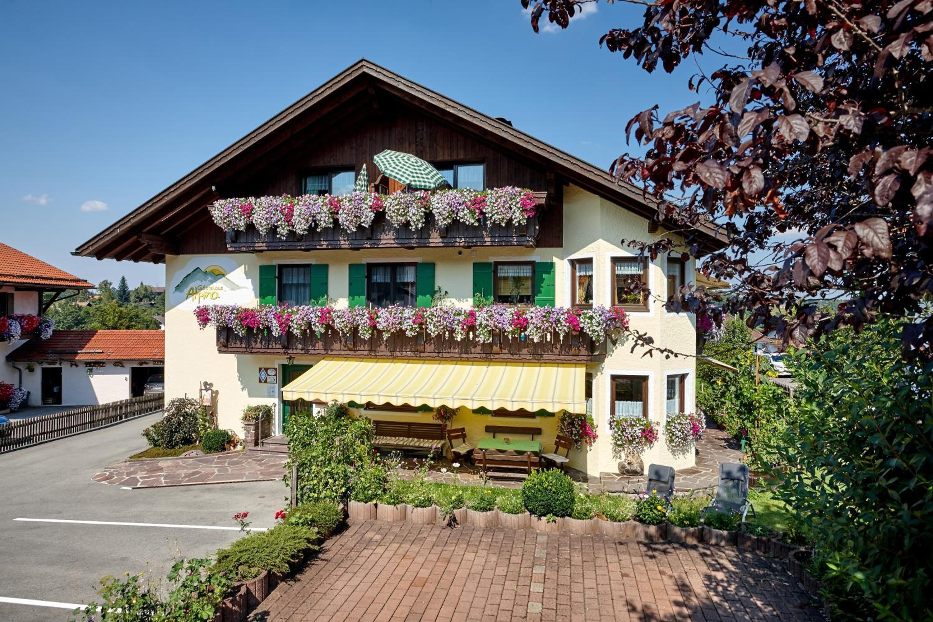 Un bâtiment de style alpin traditionnel, doté d’un balcon orné de fleurs, d’un auvent jaune et d’une cour pavée avec des sièges extérieurs.