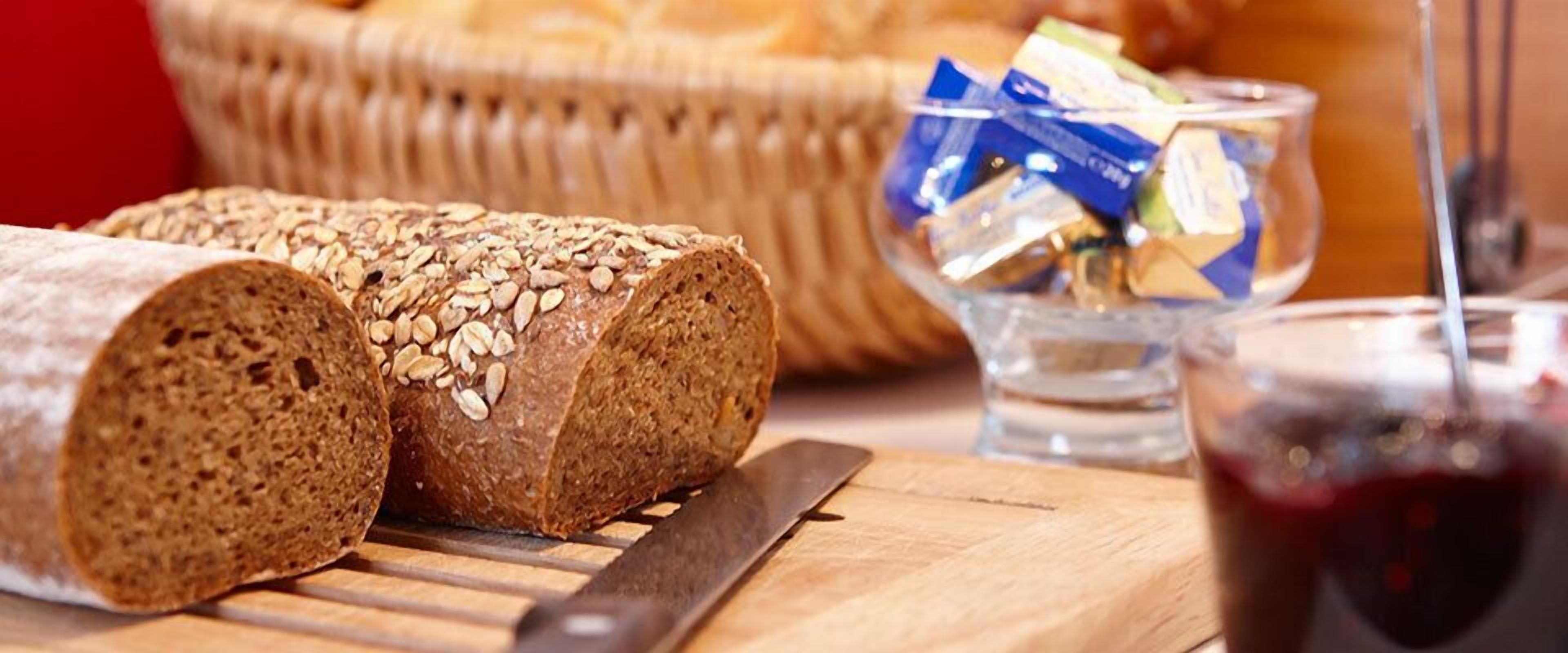Ein Roggenbrot mit Haferflocken auf einem Holzschneidebrett, ein Messer, ein Glas rote Flüssigkeit und ein Korb mit Brot im Hintergrund.