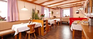 A restaurant with wooden tables and benches, white tablecloths, and hanging pendant lights. - Gästehaus Alpina (Bad Kohlgrub)