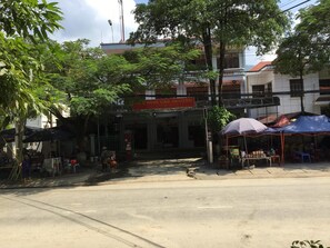Escena de carrer amb un edifici que té un rètol que diu 'Nguyen Cao Nguyen'. Hi ha arbres, un venedor ambulant amb una taula i algunes persones al fons.