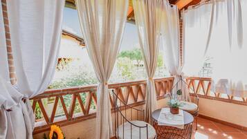 Ein Balkon mit einem Korbstuhl, einem kleinen runden Tisch mit Blumen und Blick auf Grünflächen und ein Gebäude im Hintergrund.