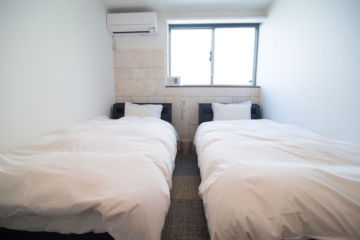 A room with two beds, a window, an air conditioning unit, and a tiled wall.