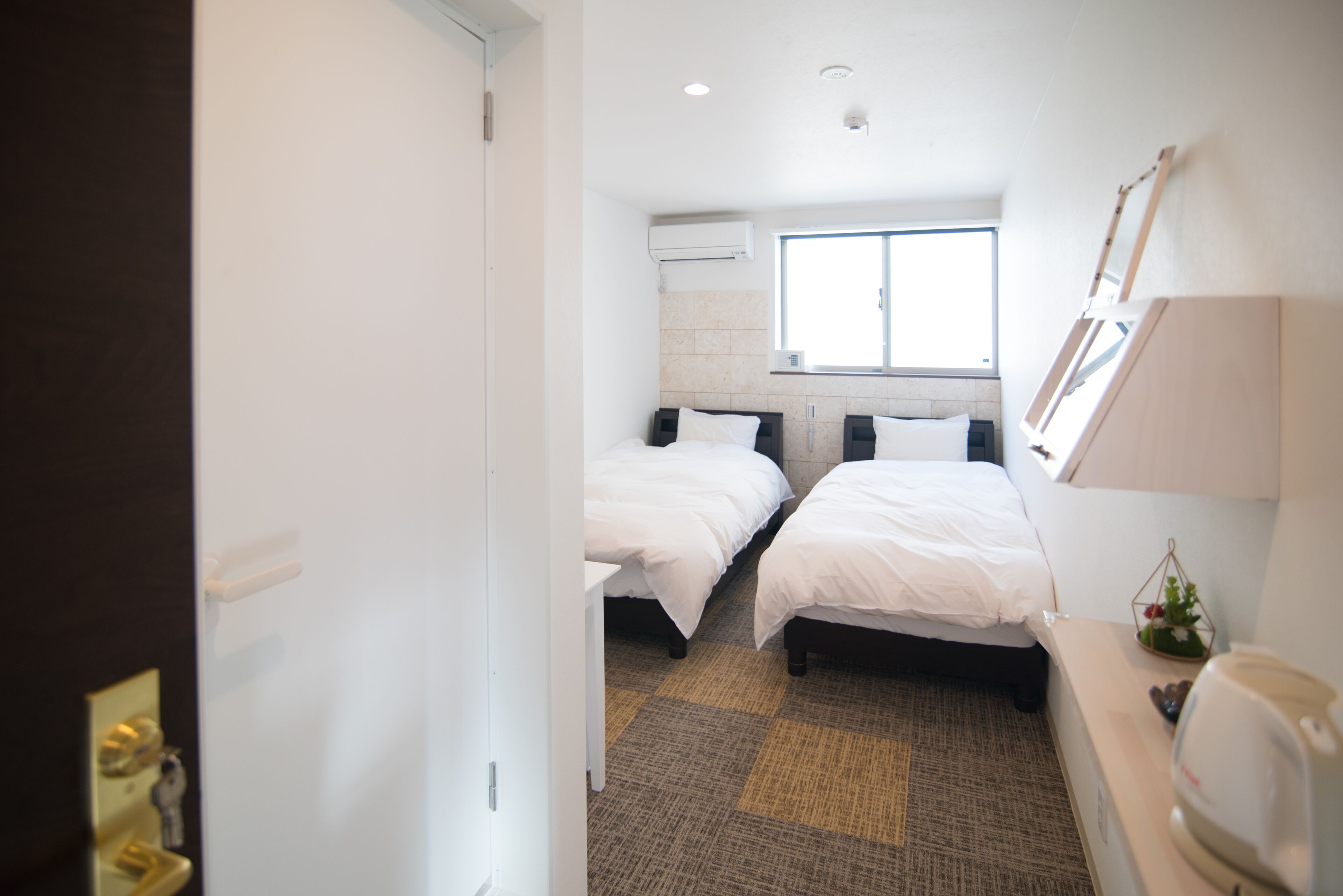 A hotel room with two beds, a window, a wall-mounted air conditioner, a small table, and a bedside lamp.