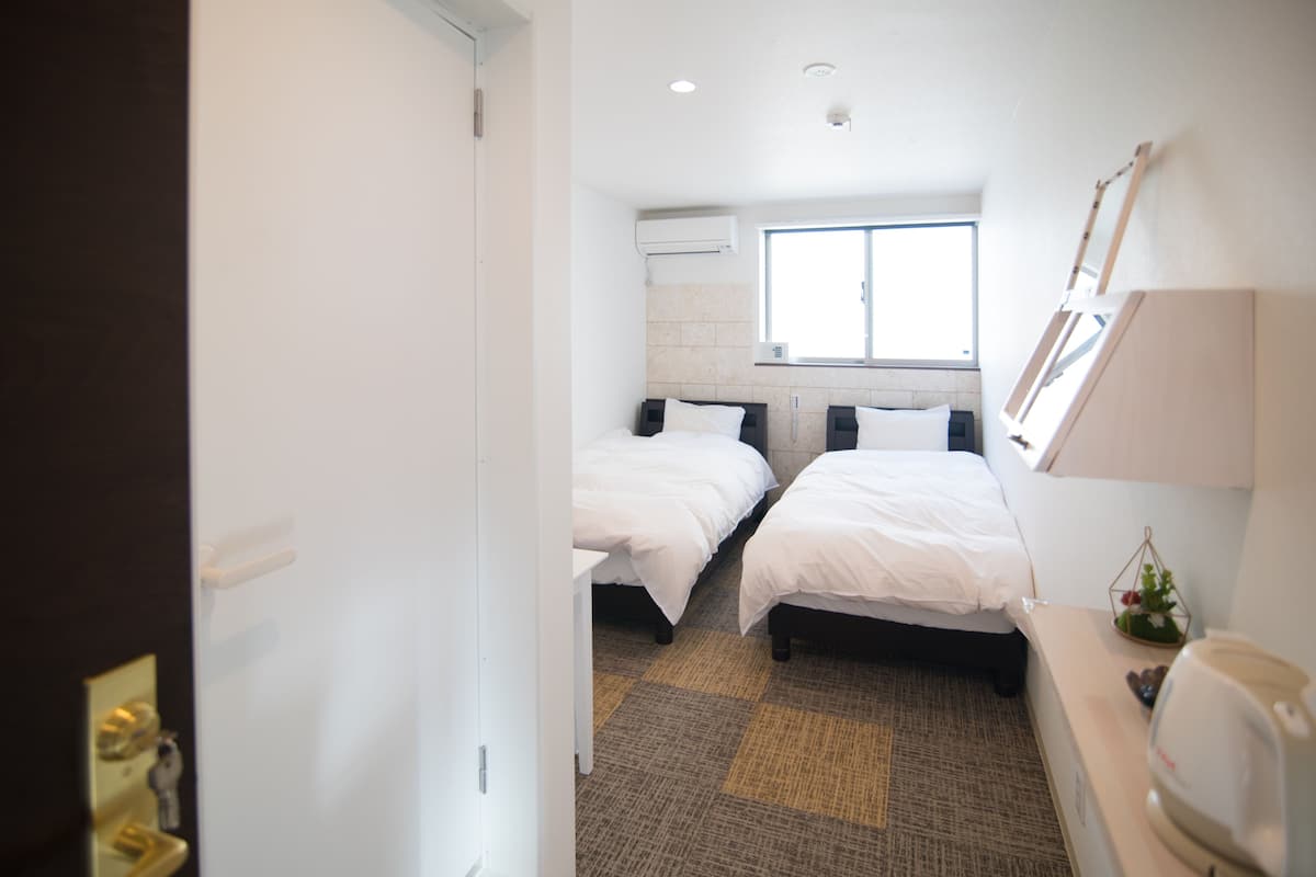 A hotel room with two beds, a window, a wall-mounted air conditioner, a small table, and a bedside lamp.