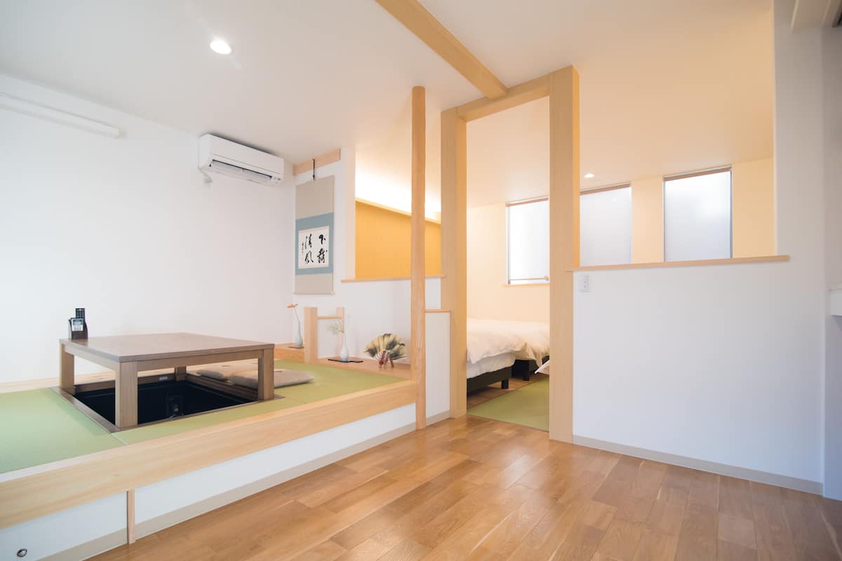 A modern, minimalist room with a wooden floor, a low table with a green mat, a bed with a white headboard, and a wall-mounted air conditioner.