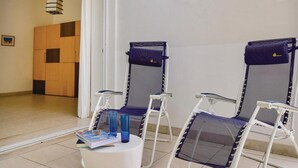 Zwei Liegestühle mit Netzgitter-Rückenlehnen und blauen Kissen, ein weißer Tisch mit einer Zeitschrift und blauen Gläsern, sowie ein Holzschrank im Hintergrund.