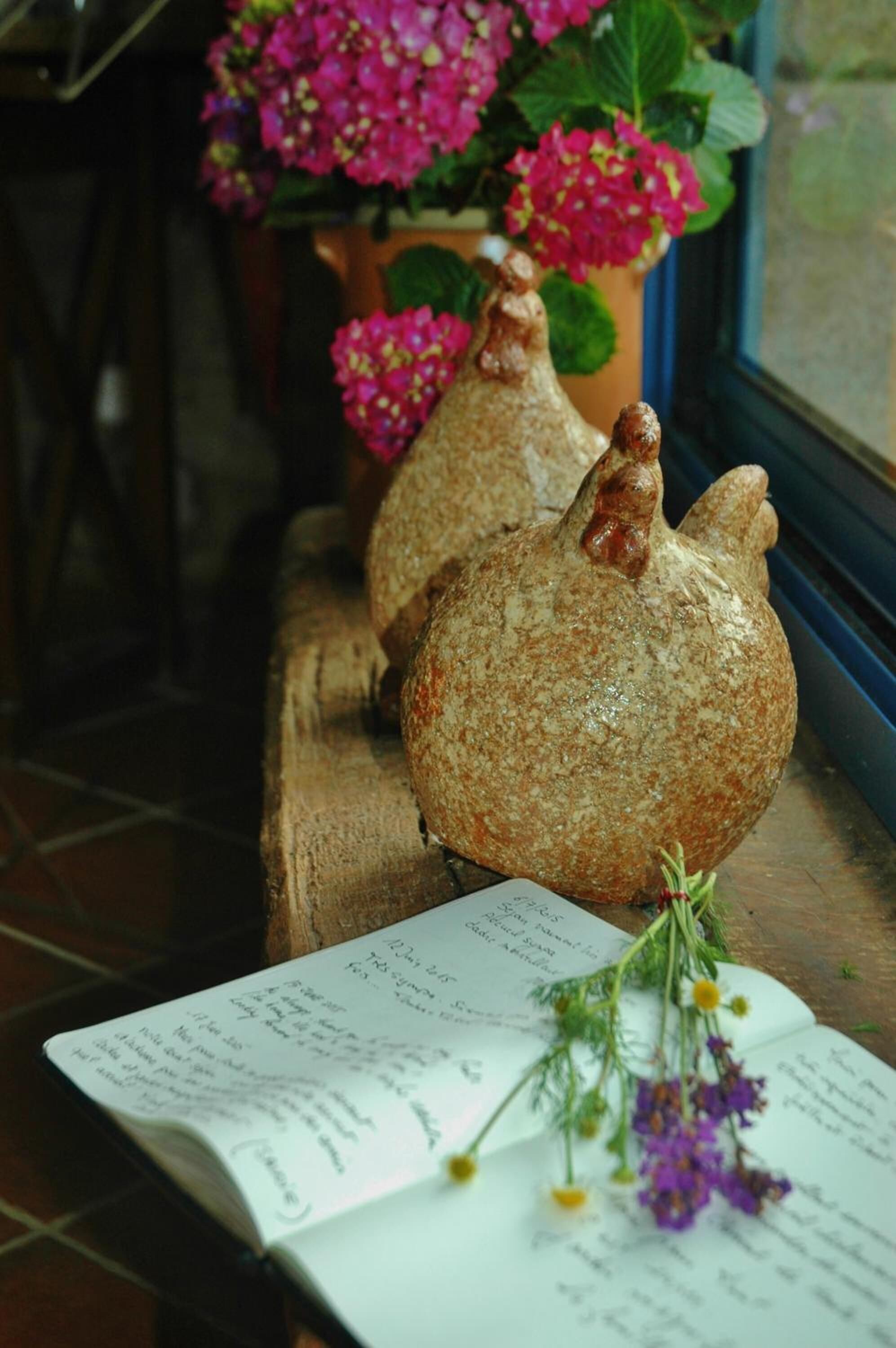 A rustic interior with ceramic chicken-shaped vases on a wooden ledge, a window with pink flowers, and a handwritten note.